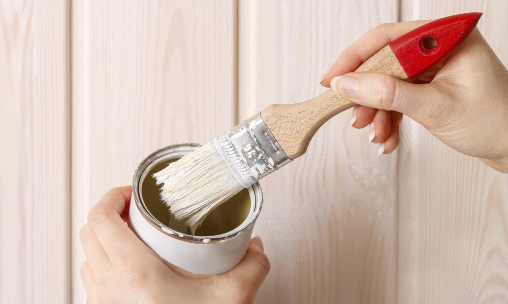 Person holding small paint can and brush in front of wood paneling