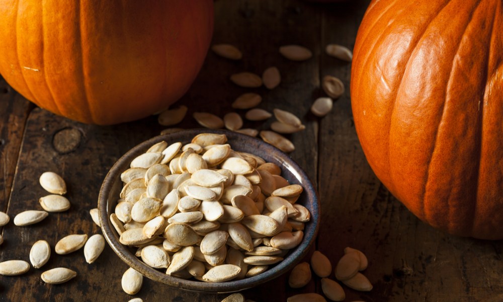 Small bowl of pumpkins seeds next to two pumpkins